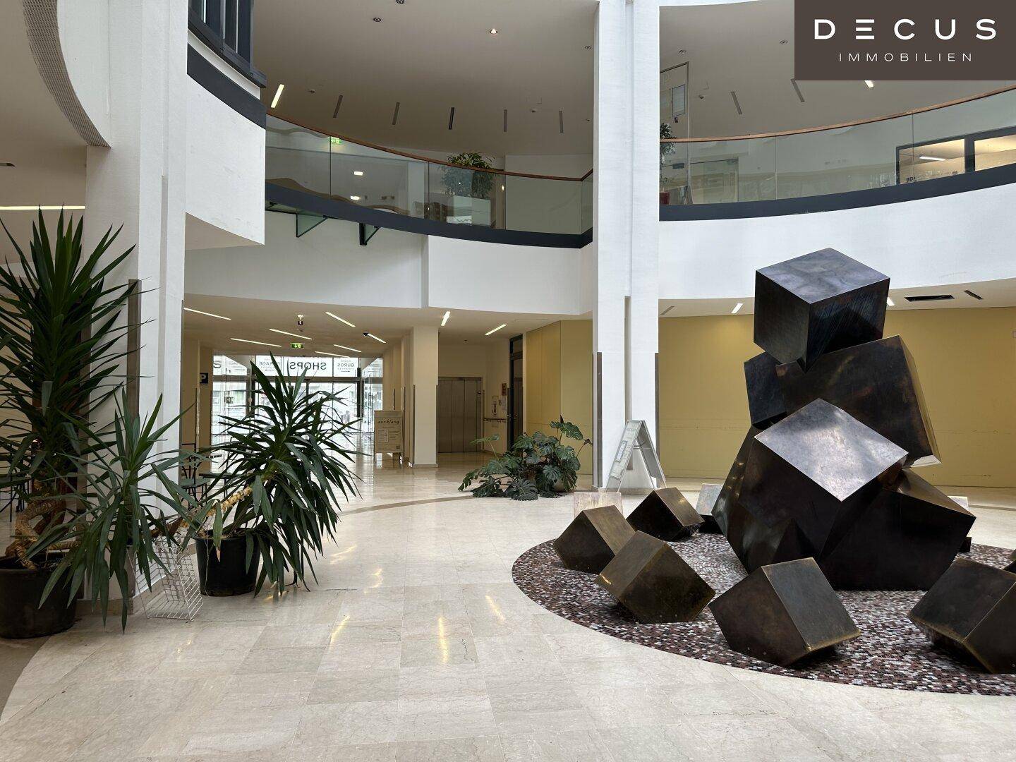 Offenes Atrium mit Pflanzen, moderner Skulptur und Blick auf die obere Etage mit Glasgeländer.