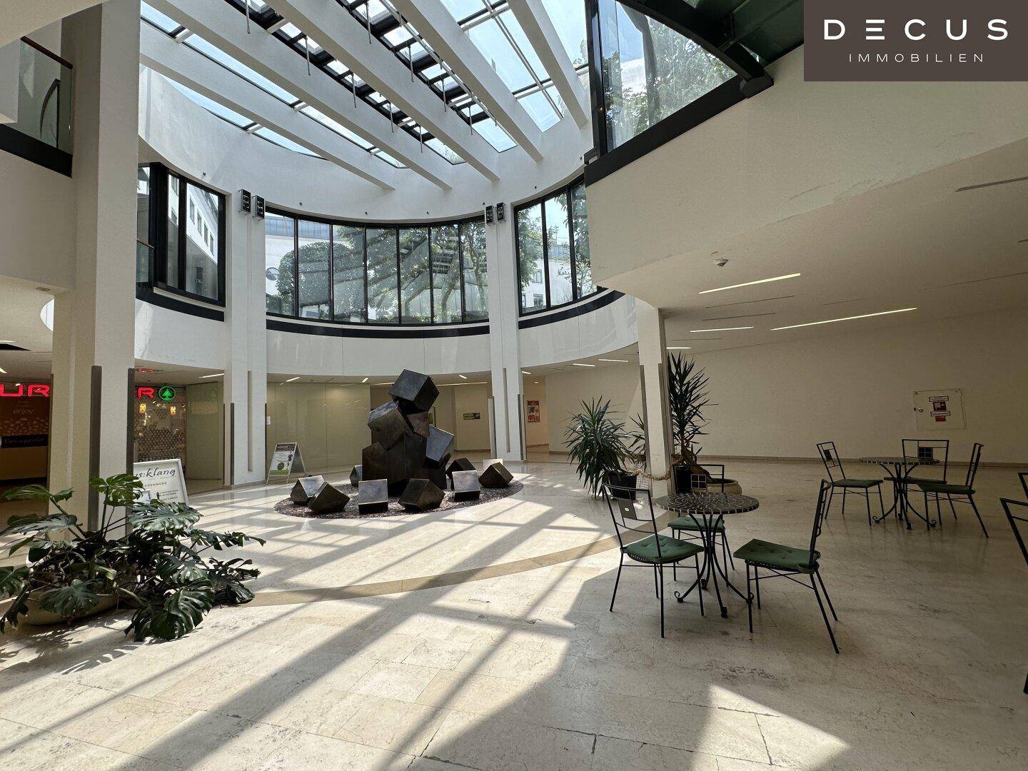 Geräumiges Atrium mit viel Tageslicht durch das Glasdach, einer Skulptur und Sitzgelegenheiten.