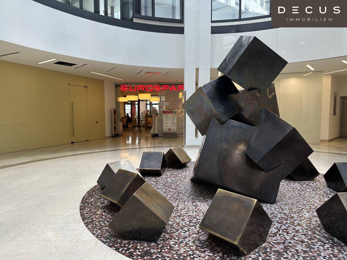Nahaufnahme einer modernen Skulptur im Atrium, mit Blick auf den Eingang eines Supermarktes.