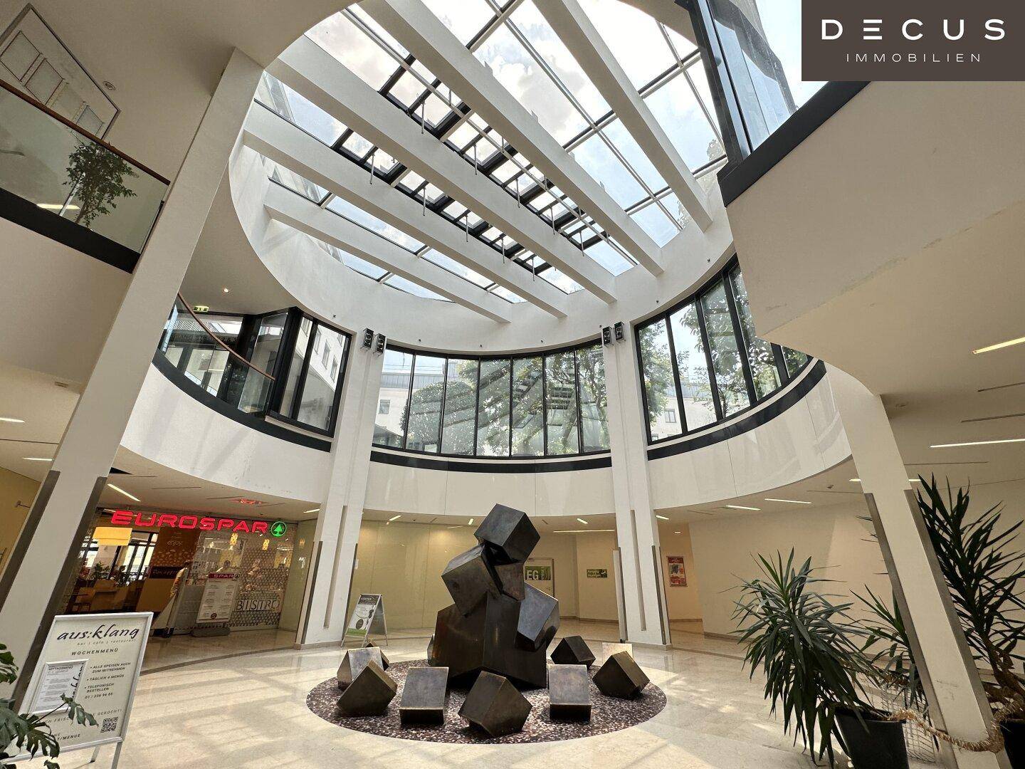 Weitläufiges Atrium mit Glasdach, moderner Skulptur und Zugang zu verschiedenen Geschäften.