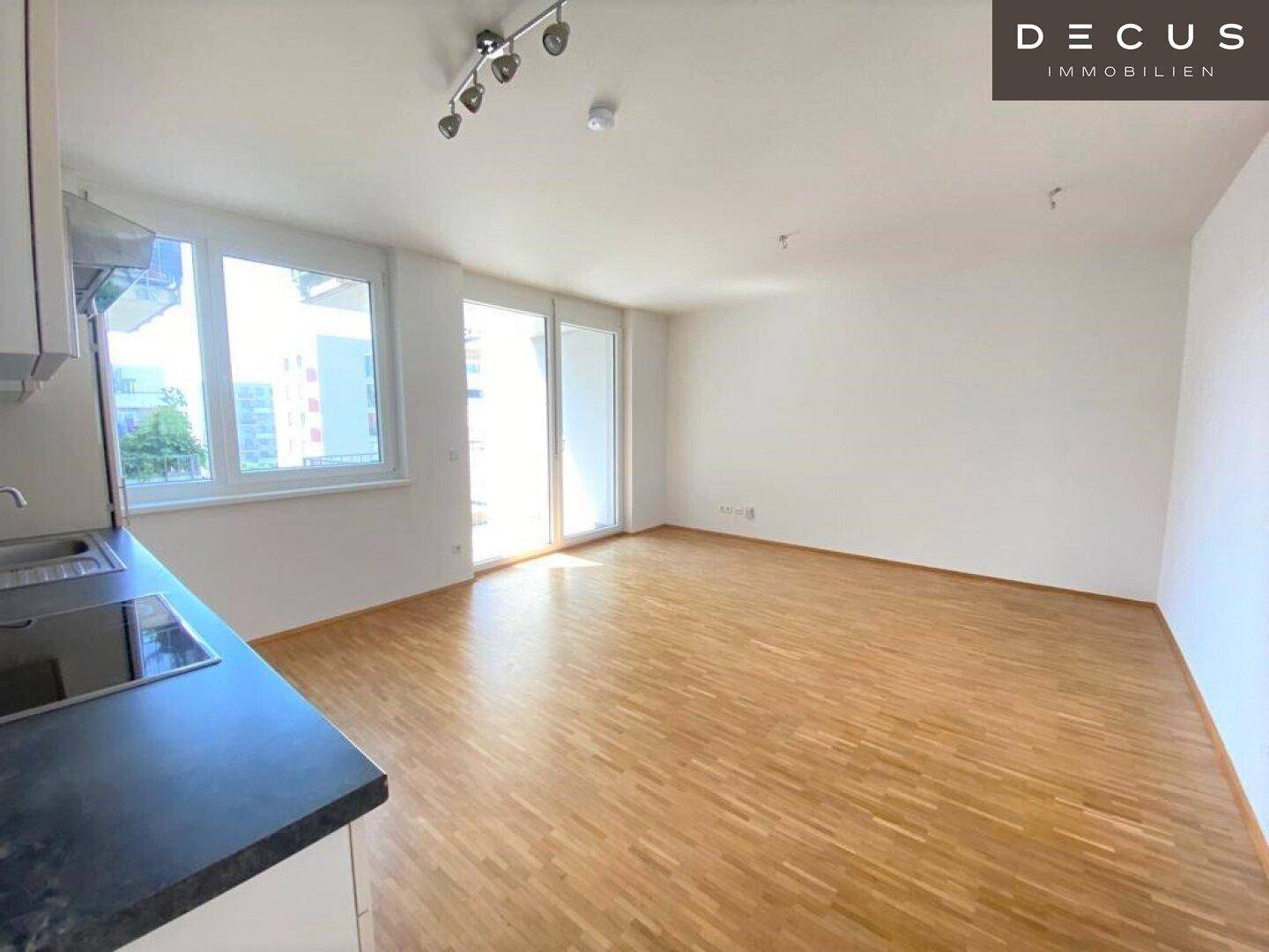 Offener Wohnbereich mit Küchenzeile, hellem Parkettboden und Zugang zu einem Balkon.