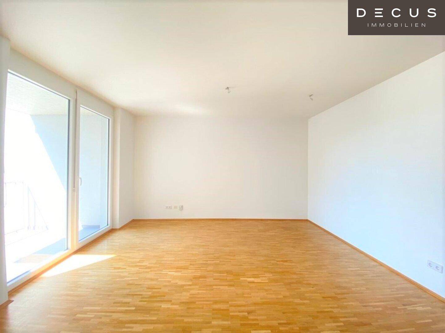 Heller, leerer Wohnraum mit Parkettboden, weißen Wänden und großen Fenstern zum Balkon.