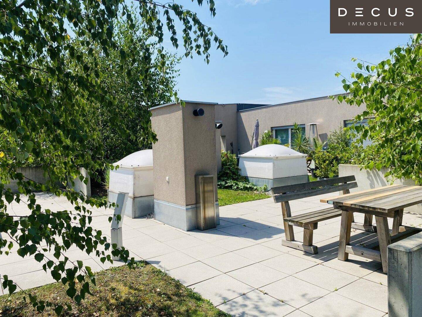 Gemeinschaftlicher Gartenbereich mit Holztisch, Bänken und viel Grün unter blauem Himmel.