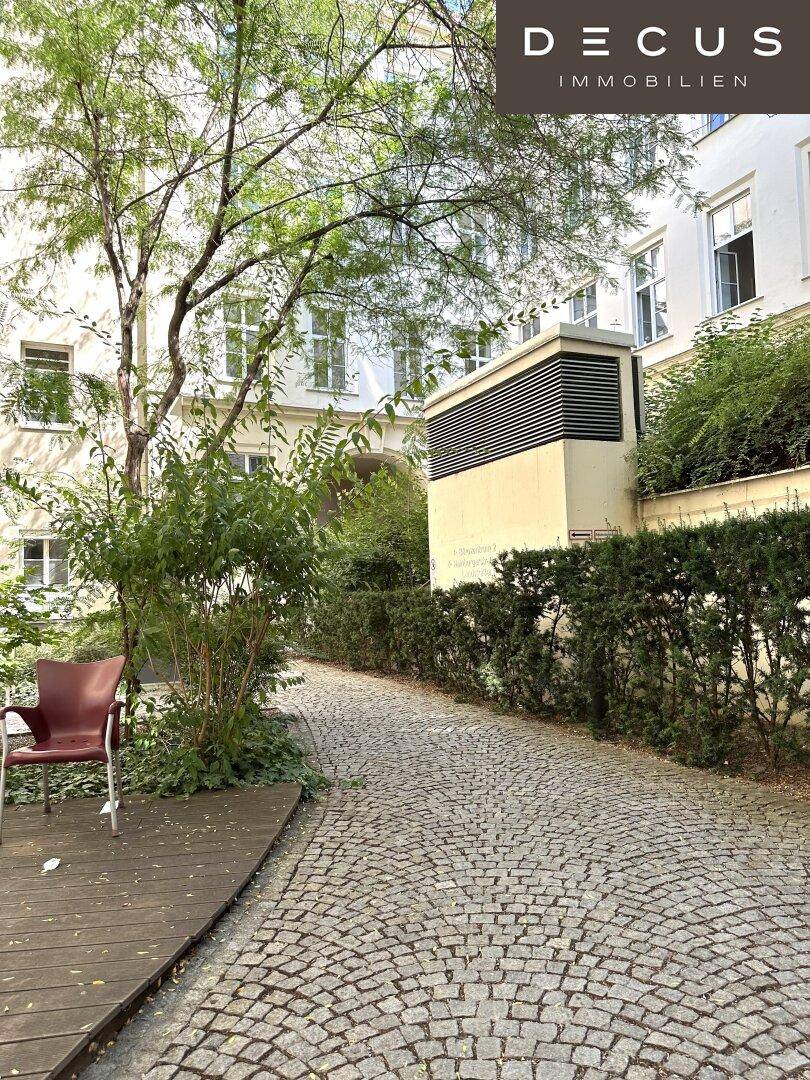 Grüner Innenhof mit Kopfsteinpflasterweg, Bäumen und einer kleinen Holzterrasse mit Sitzgelegenheit.