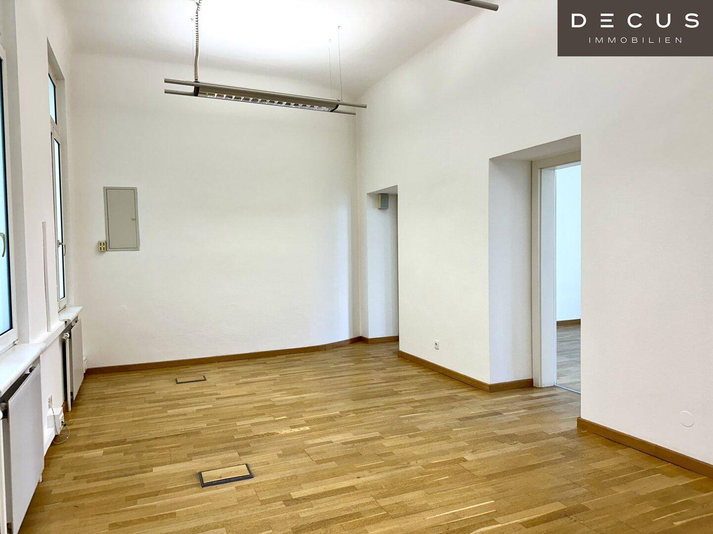 Geräumiger, leerer Büroraum mit Holzboden, weißen Wänden und zwei Fenstern, ausgestattet mit Deckenleuchten.