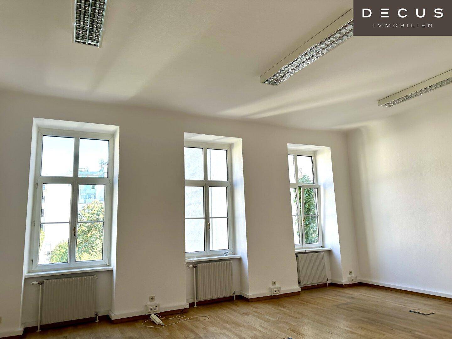 Heller Büroraum mit Holzboden, weißen Wänden und drei hohen Fenstern, die viel Tageslicht bieten.