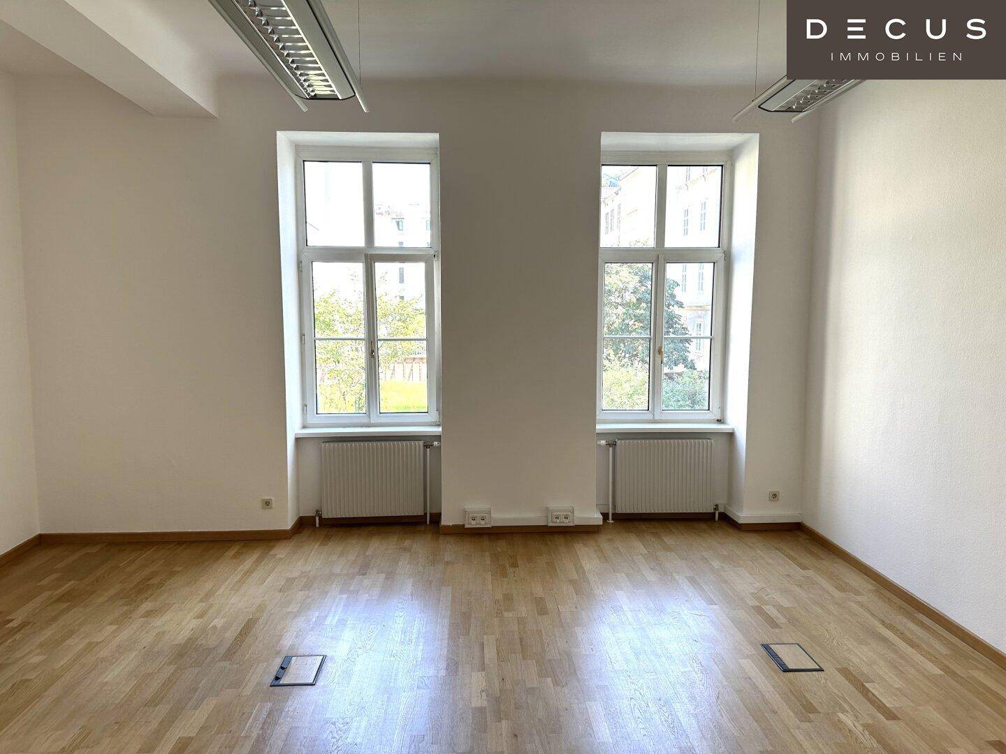Heller Büroraum mit zwei großen Fenstern und Holzboden, bietet eine angenehme Arbeitsatmosphäre.