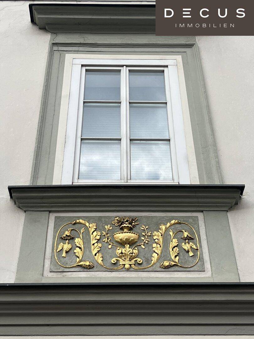 Detailansicht einer Fensterfront mit aufwendiger goldener Stuckverzierung unter dem Fenster.