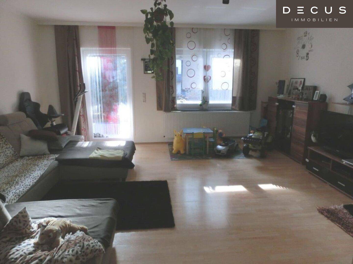 Geräumiges Wohnzimmer mit hellem Laminatboden, großem Sofa und viel Tageslicht.