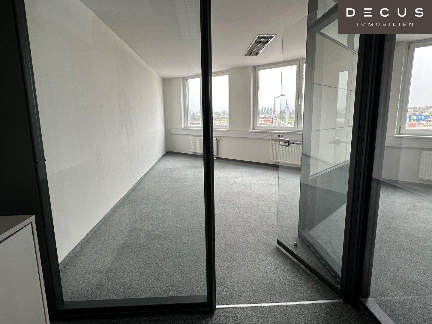Blick in ein helles Büro mit Teppichboden und großen Fenstern, die viel Tageslicht hereinlassen.