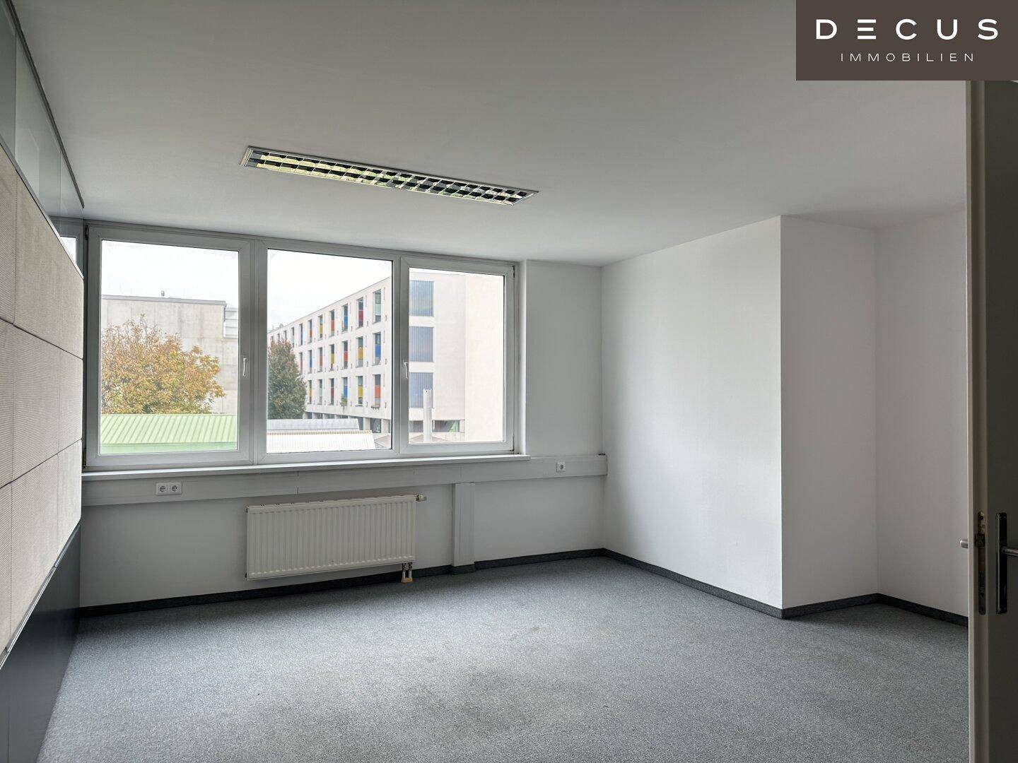 Helles Büro mit Teppichboden und Blick auf umliegende Gebäude durch große Fenster.