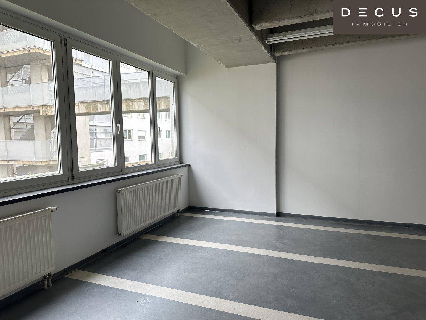 Raum mit großen Fenstern, die den Blick auf ein angrenzendes Gebäude freigeben, Heizkörpern und gestreiftem Fliesenboden.