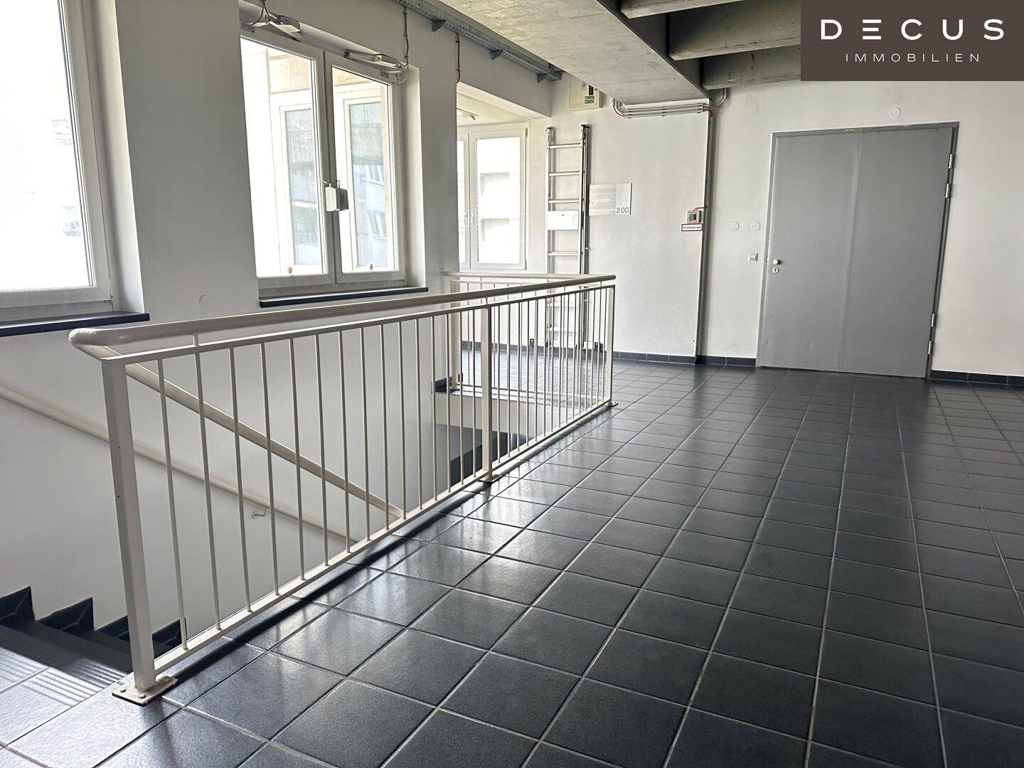 Heller Treppenhausbereich mit Metallgeländer und dunklen Fliesen, der zu weiteren Etagen führt.