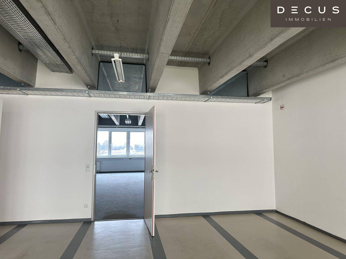 Flur mit Zugang zu einem hellen Büroraum, gekennzeichnet durch sichtbare Deckenbalken.