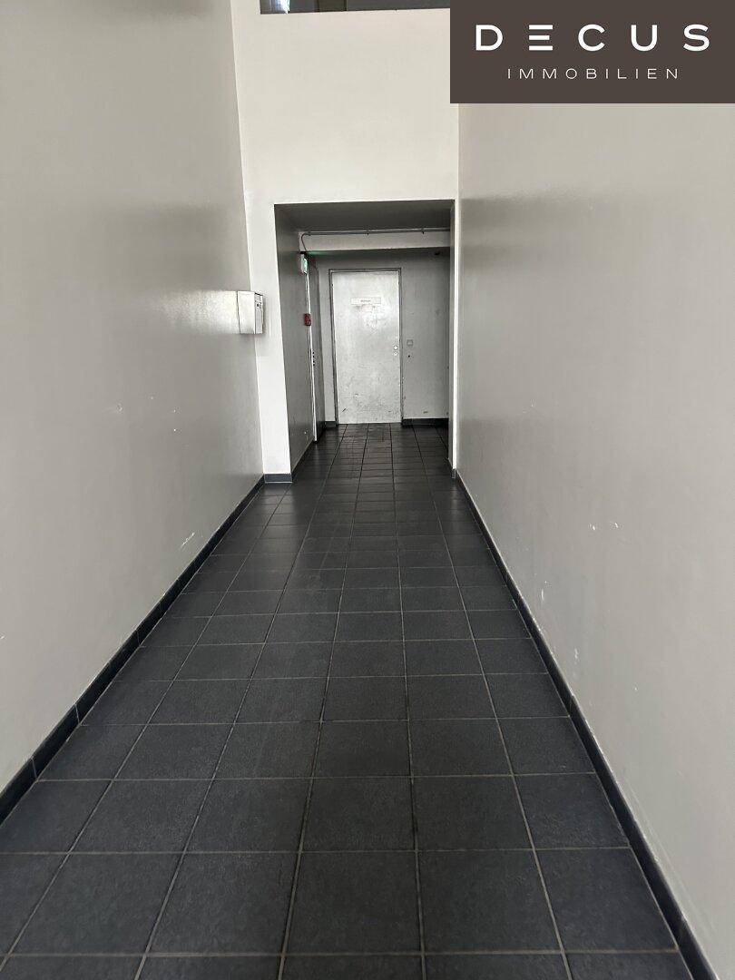 Schmaler Flur mit dunklem Fliesenboden und weißen Wänden, der zu einer Tür am Ende führt.