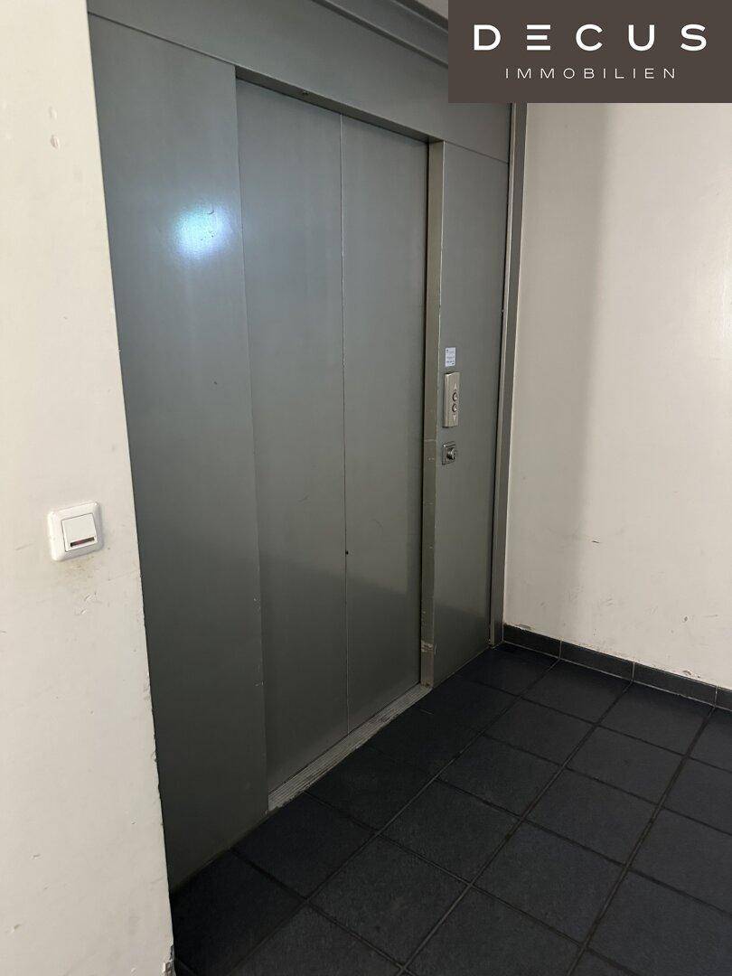 Graue Aufzugstür mit Rufknopf und dunklem Fliesenboden im Eingangsbereich.