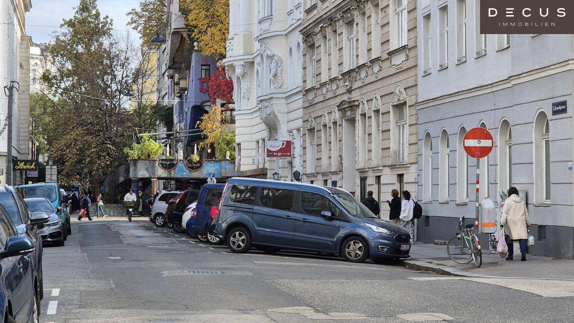 Belebte Straße mit geparkten Autos und Fußgängern in einem städtischen Viertel mit Altbauten.