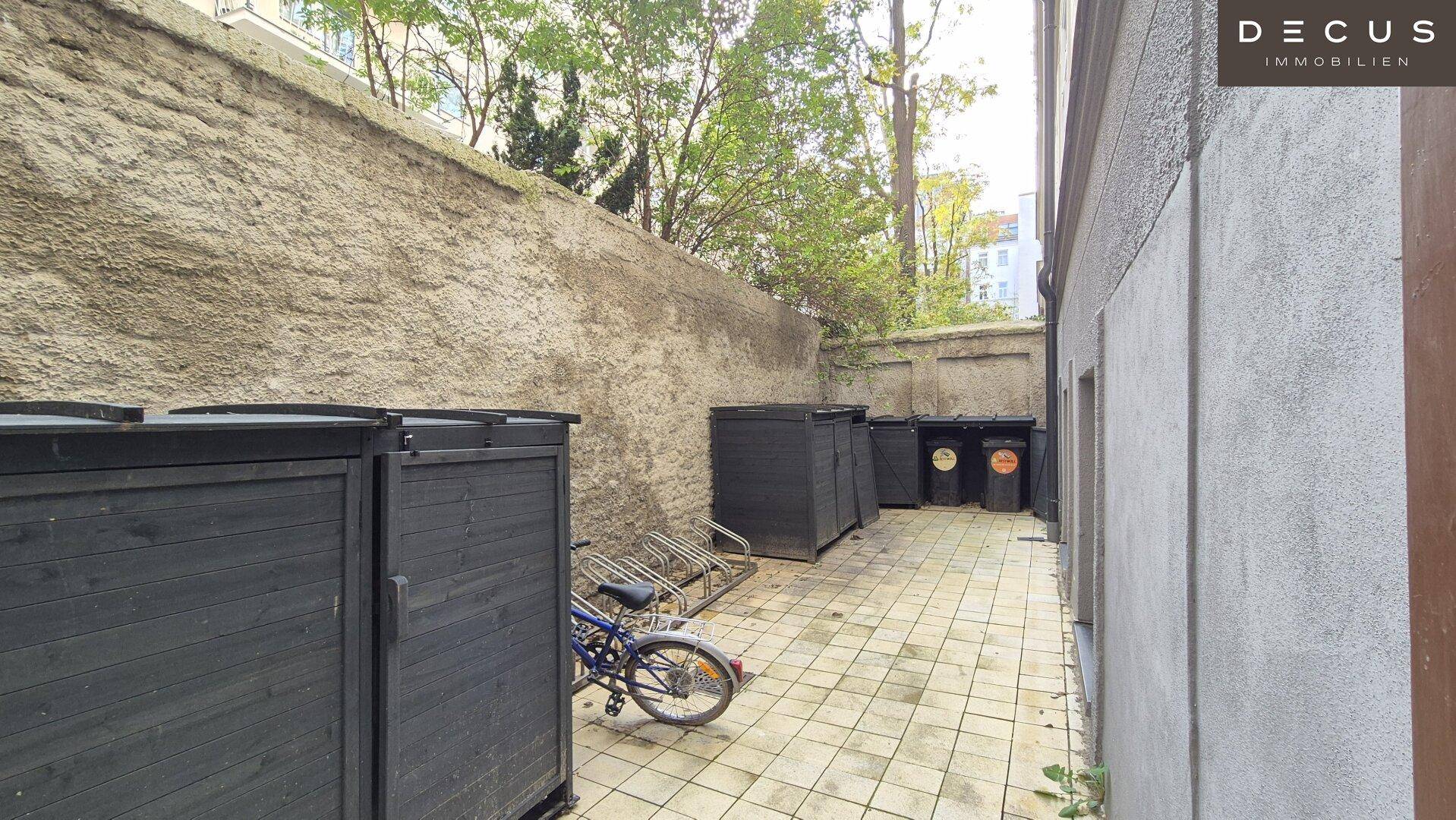 Hinterhof mit Fahrradständern, Mülltonnen und einer hohen Mauer, dient als Abstellfläche.