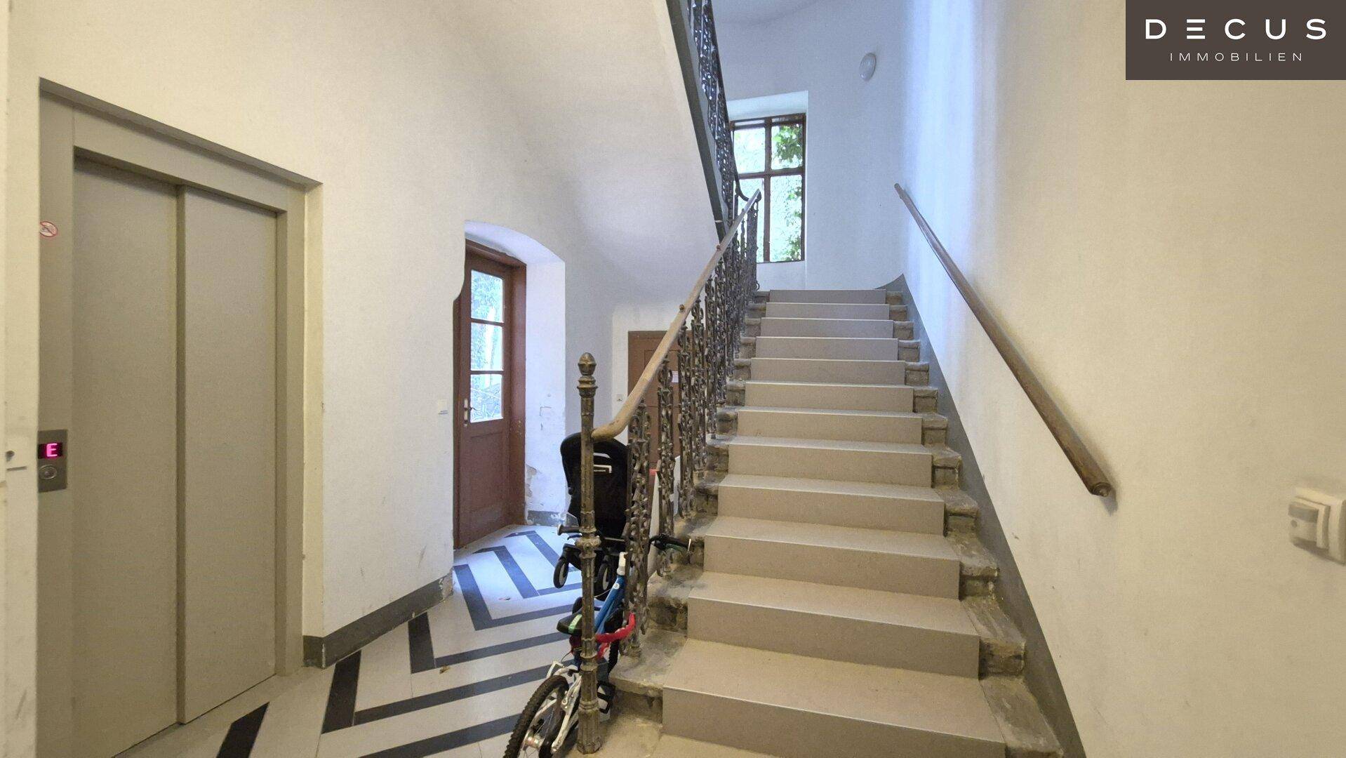 Historisches Treppenhaus mit schmiedeeisernem Geländer, Aufzug und natürlichem Lichteinfall durch ein Fenster.