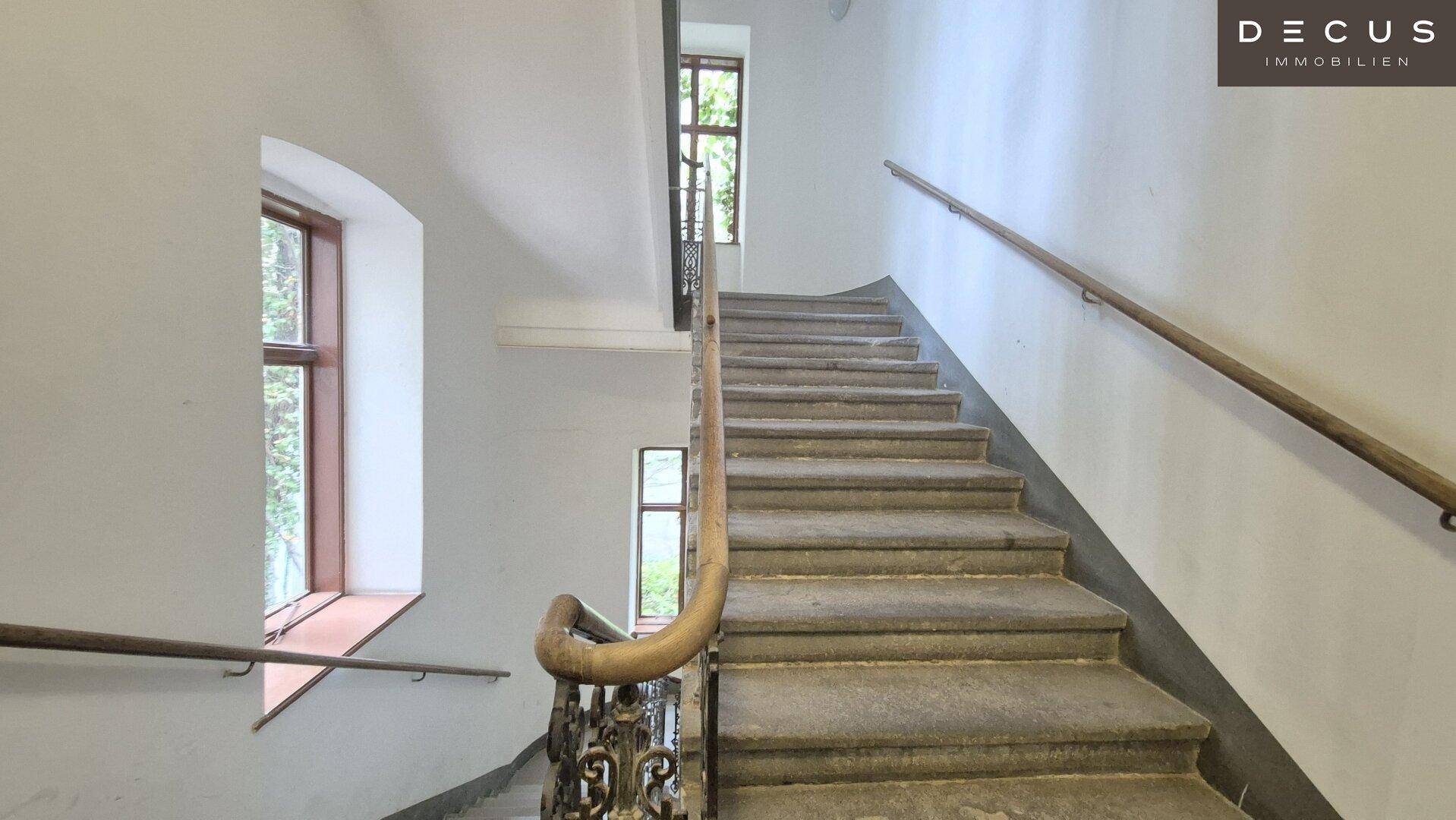 Breites Treppenhaus mit Steinstufen, Holzhandlauf und Fenstern, die natürliches Licht hereinlassen.