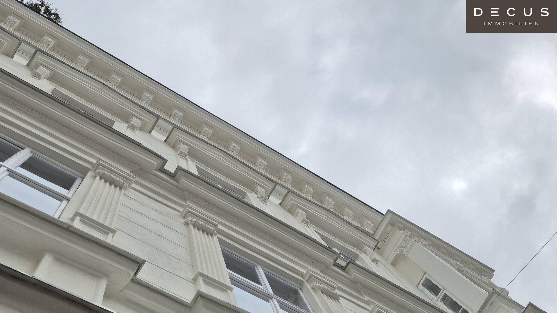Obere Fassade eines historischen Gebäudes mit detaillierten Stuckverzierungen unter einem bewölkten Himmel.