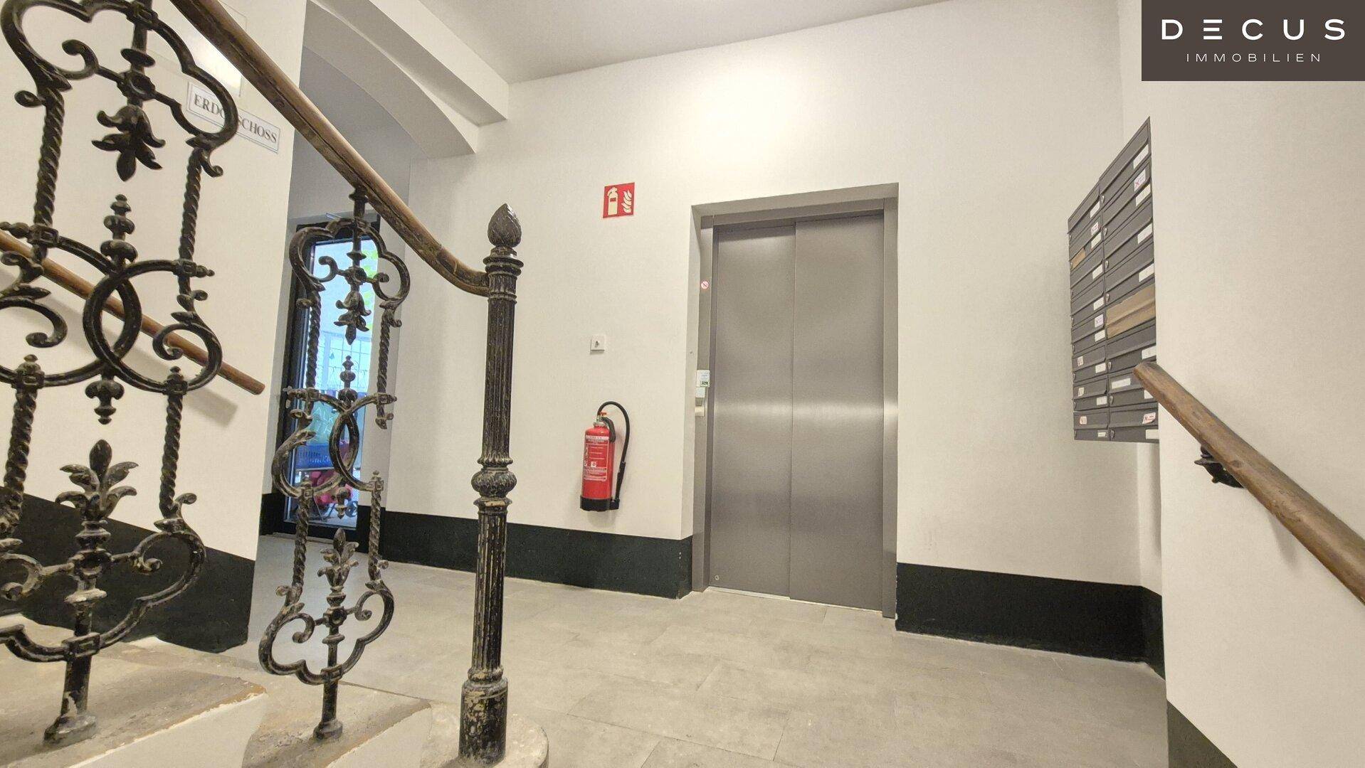 Heller Hausflur mit historischem Treppengeländer und modernem Aufzug für bequemen Zugang zu den Etagen.