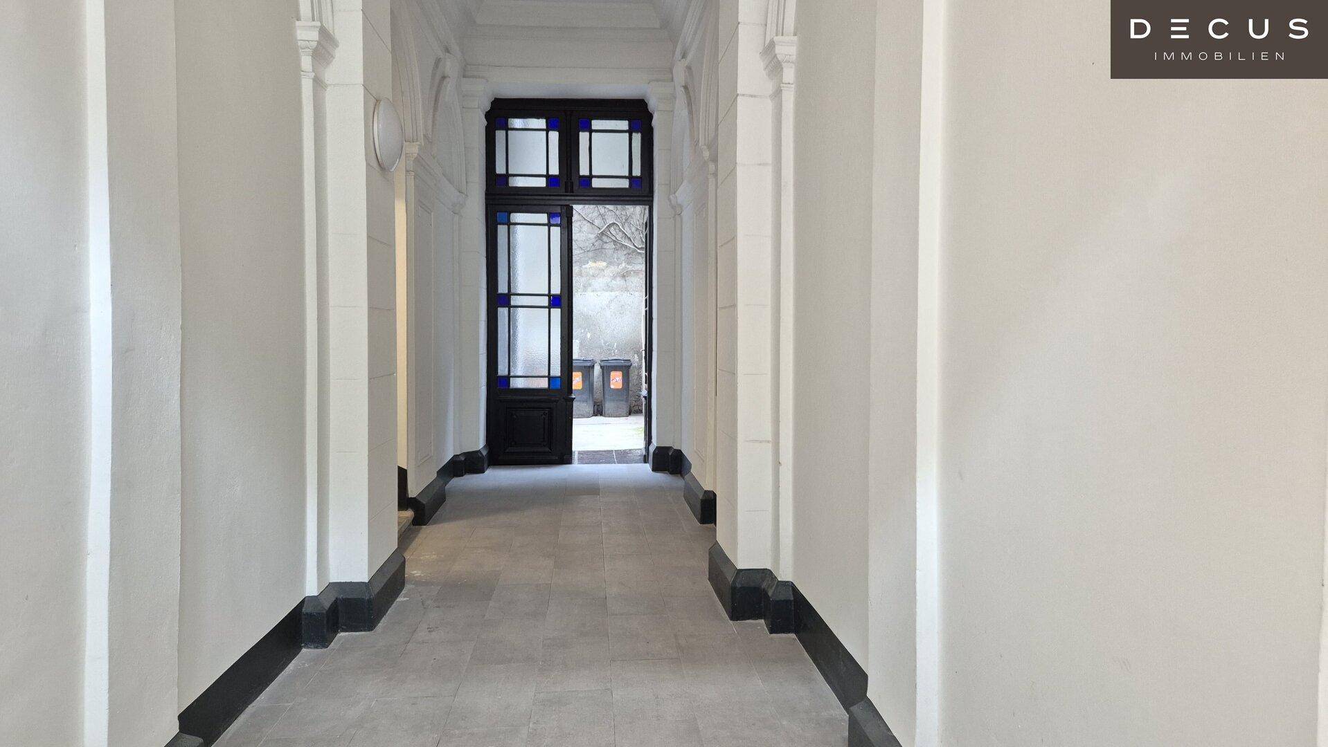 Langer, heller Flur mit Fliesenboden und hohen Decken, der zum Ausgang des Gebäudes führt.