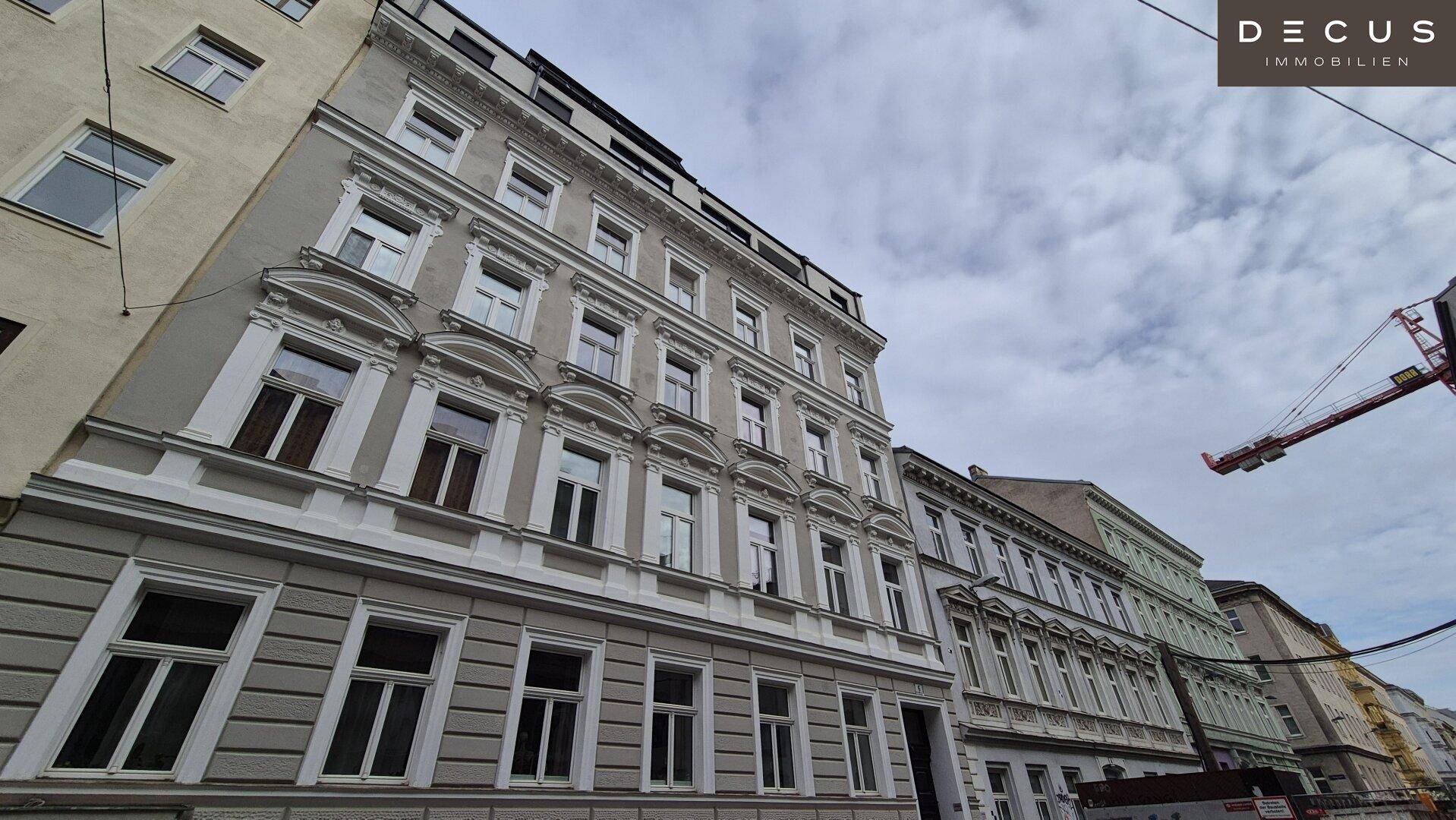 Weitwinkelansicht der historischen Gebäudefassade mit modernen Dachaufbauten unter bewölktem Himmel.