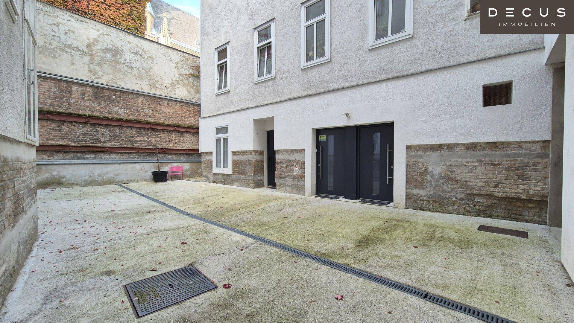 Betonierter Innenhof mit Entwässerungsrinnen und zwei modernen schwarzen Eingangstüren im Erdgeschoss.