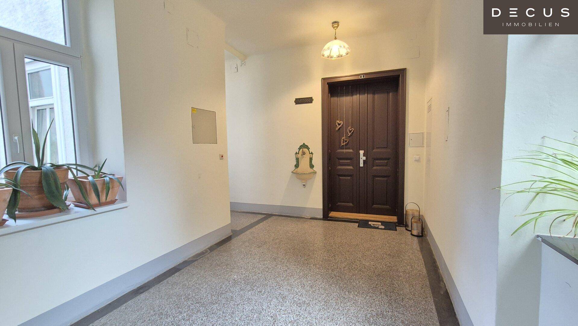 Heller Flur auf einer Etage mit Terrazzoboden, Pflanzen und einer dunklen Wohnungstür.