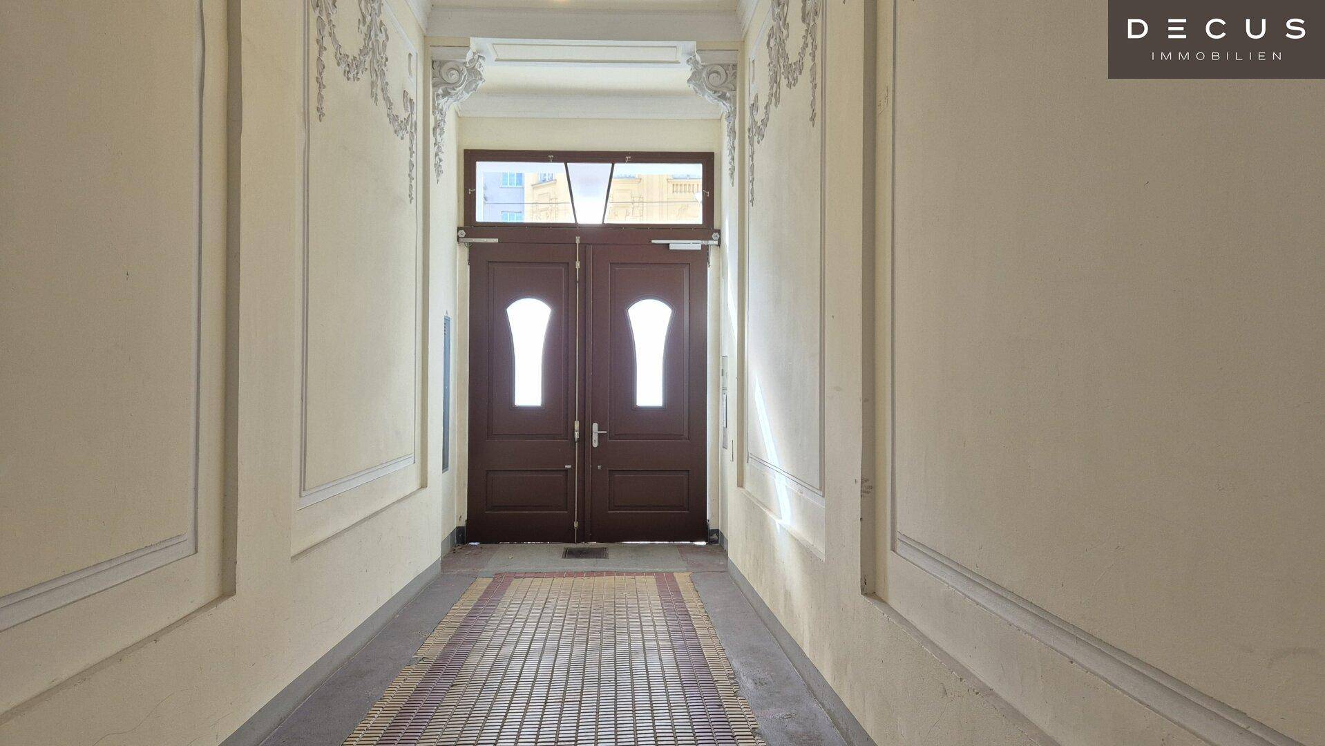 Langer, heller Flur mit gemustertem Boden und verzierten Wänden, der zum Haupteingang führt.