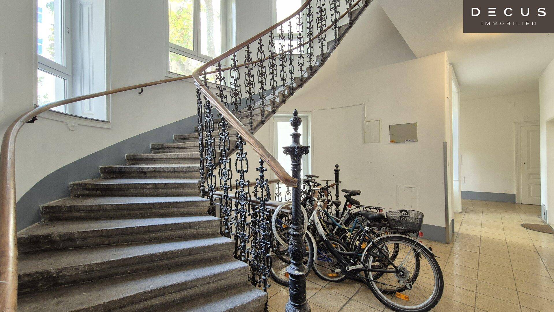 Historisches Treppenhaus mit schmiedeeisernem Geländer und Steinstufen, Fahrräder im Eingangsbereich abgestellt.