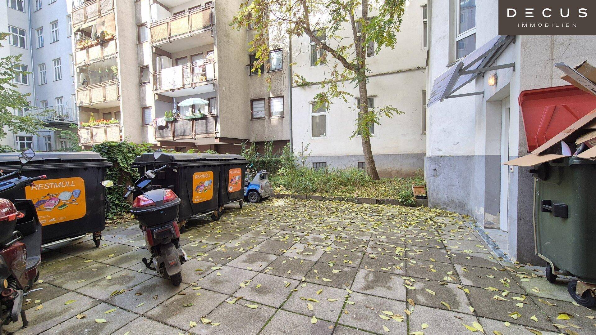 Gepflasterter Innenhof mit Mülltonnen und abgestellten Rollern, umgeben von Gebäuden und Bäumen.