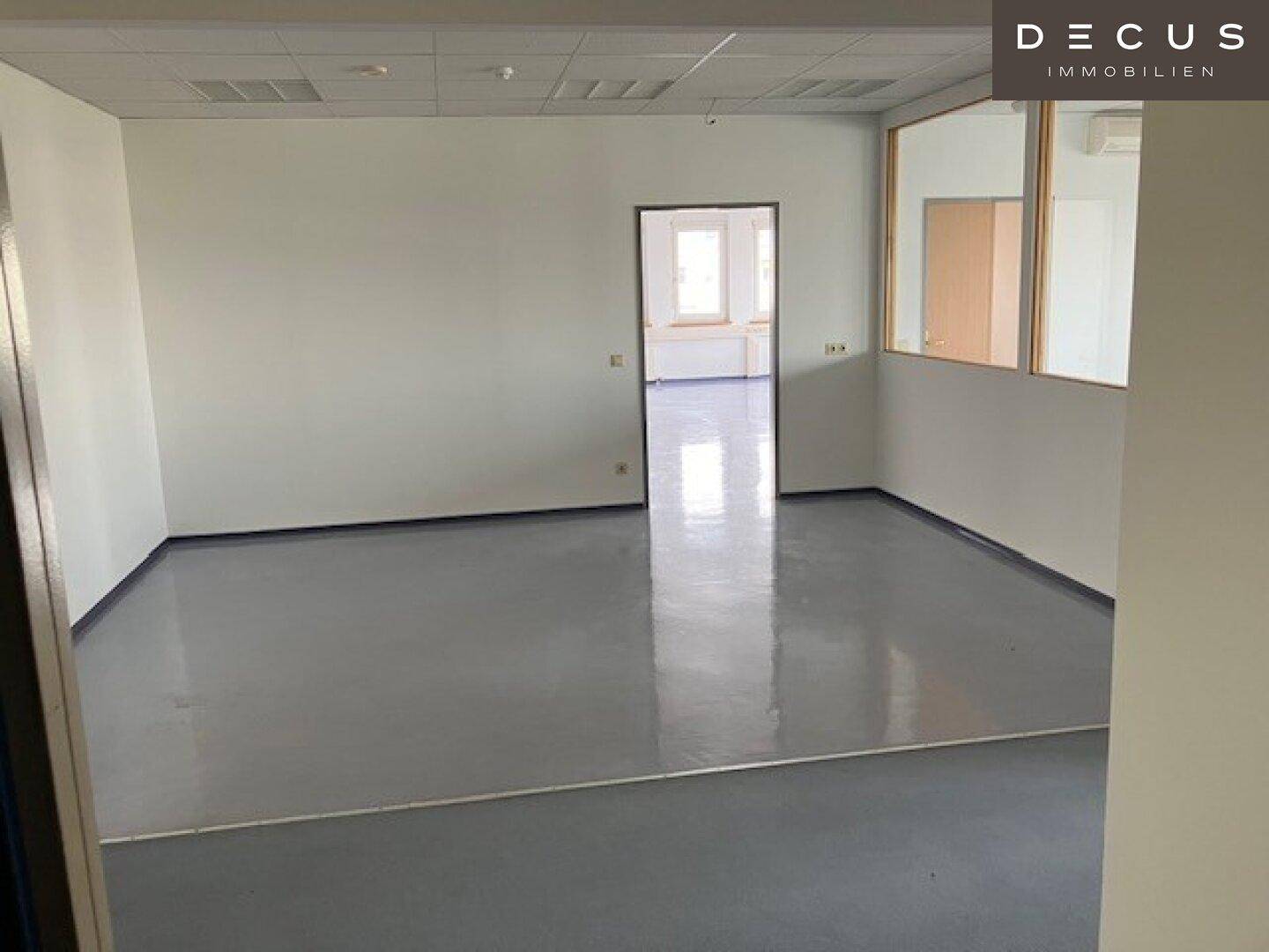 Leeres Büro mit hellgrauem Vinylboden, weißen Wänden und Blick in einen weiteren angrenzenden Raum.