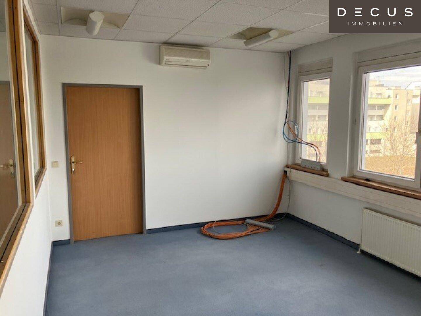 Leeres Büro mit blauer Teppichboden, weißen Wänden, zwei Fenstern und einer Klimaanlage.