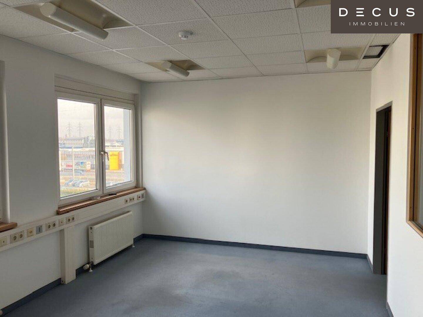 Leeres Büro mit blau-grauem Teppichboden, großen Fenstern und Heizkörpern unter der Fensterbank.
