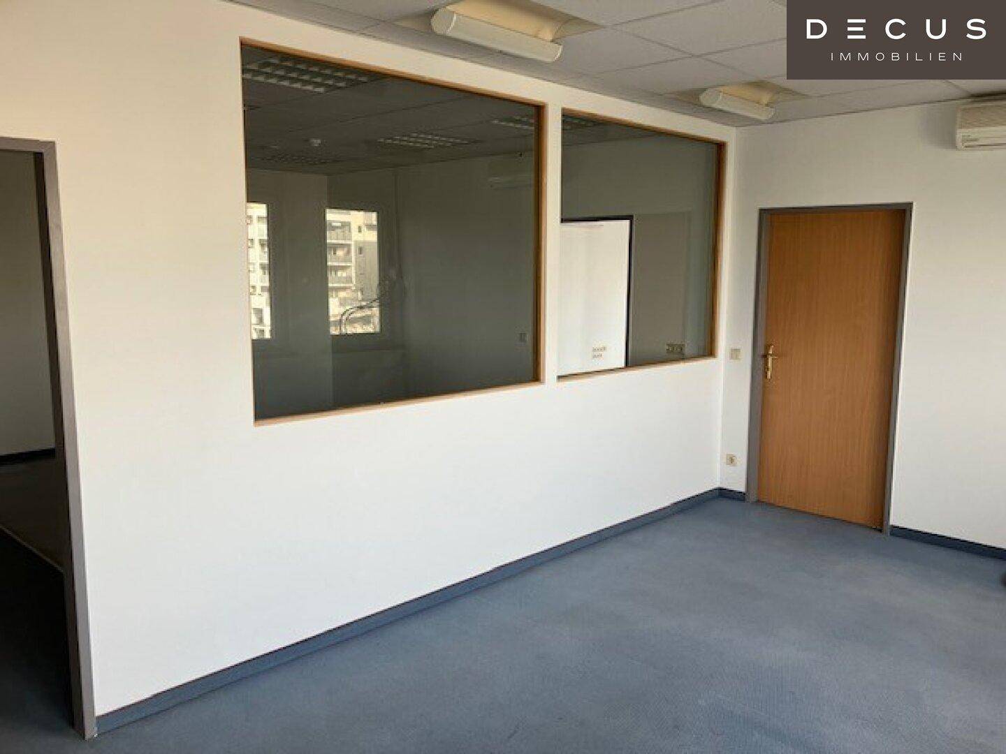 Leeres Büro mit blau-grauem Teppichboden, Holztür und Glaswänden zu einem angrenzenden Raum.