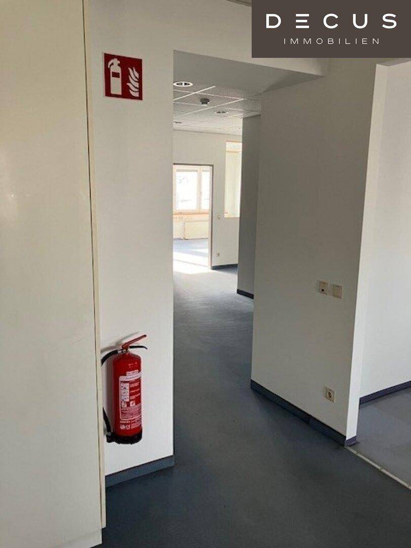 Langer Flur mit Feuerlöscher, hellgrauem Vinylboden und Türen zu verschiedenen Büroräumen.