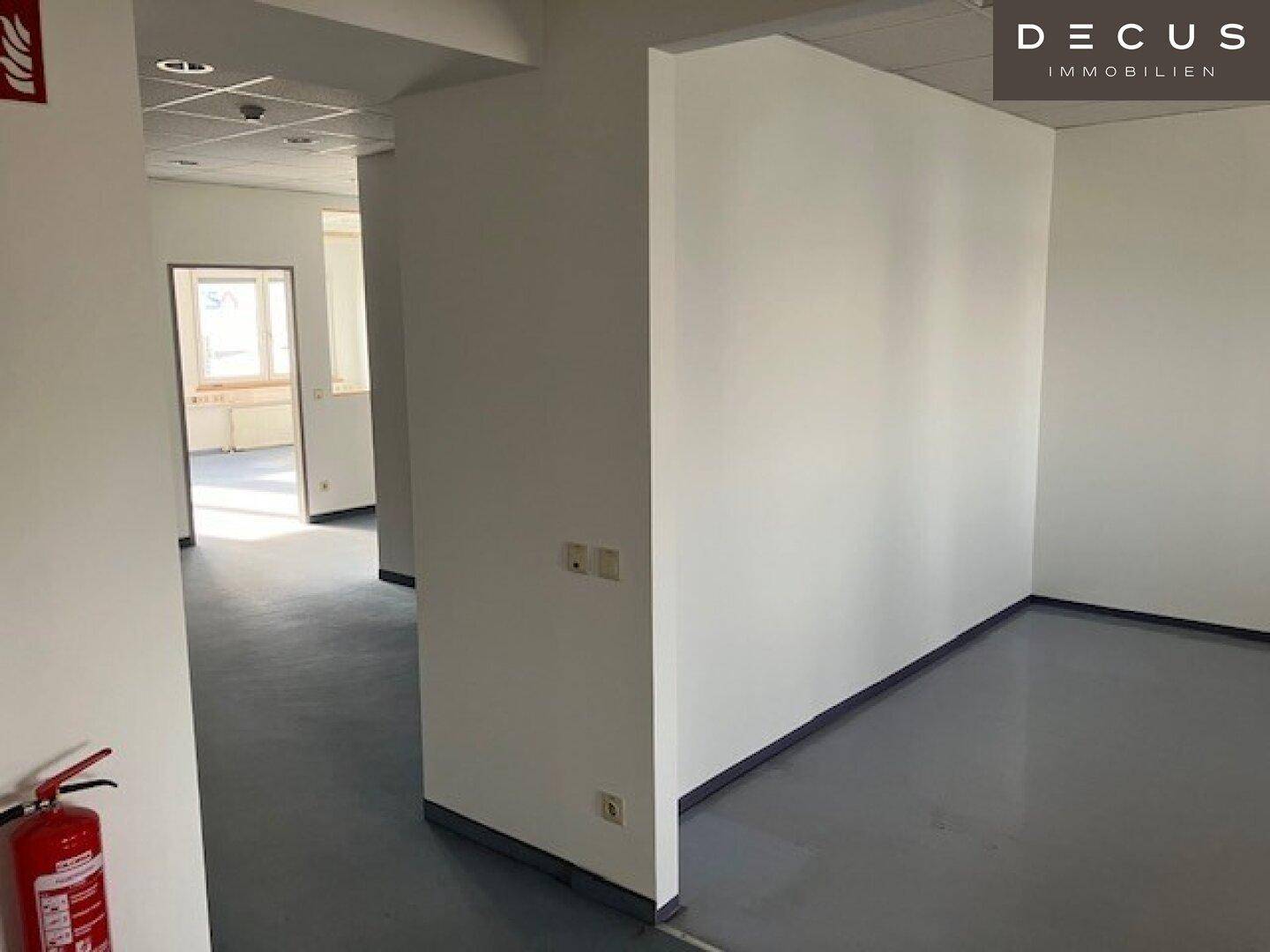 Leerer Büroraum mit hellgrauem Vinylboden, weißen Wänden und einem Feuerlöscher im Vordergrund.
