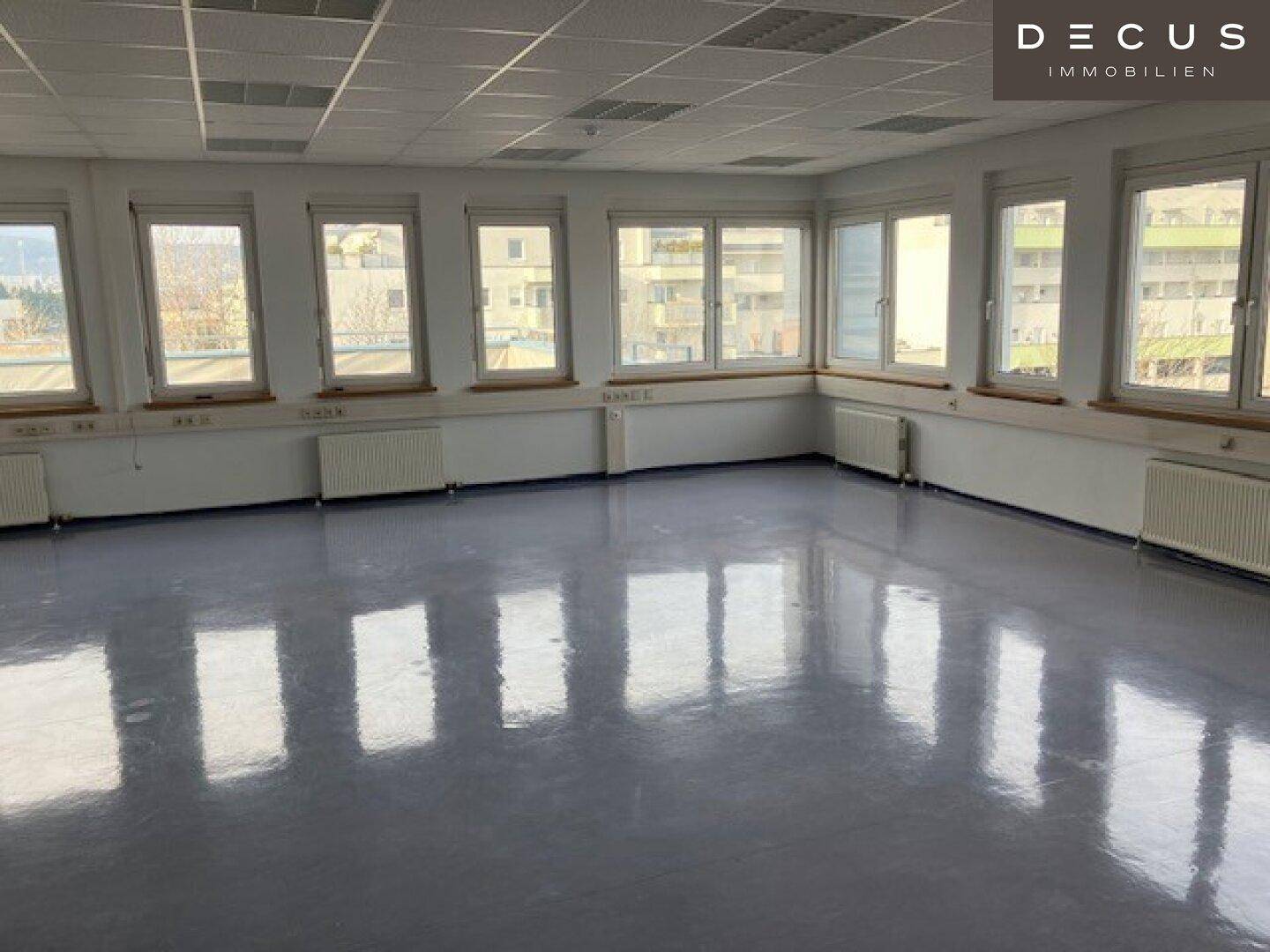 Großes, leeres Büro mit vielen Fenstern, hellgrauem Vinylboden und Heizkörpern.
