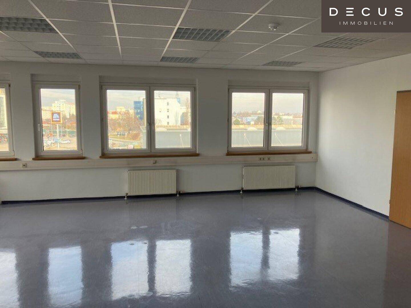 Großes, leeres Büro mit vielen Fenstern, hellgrauem Vinylboden und Heizkörpern.