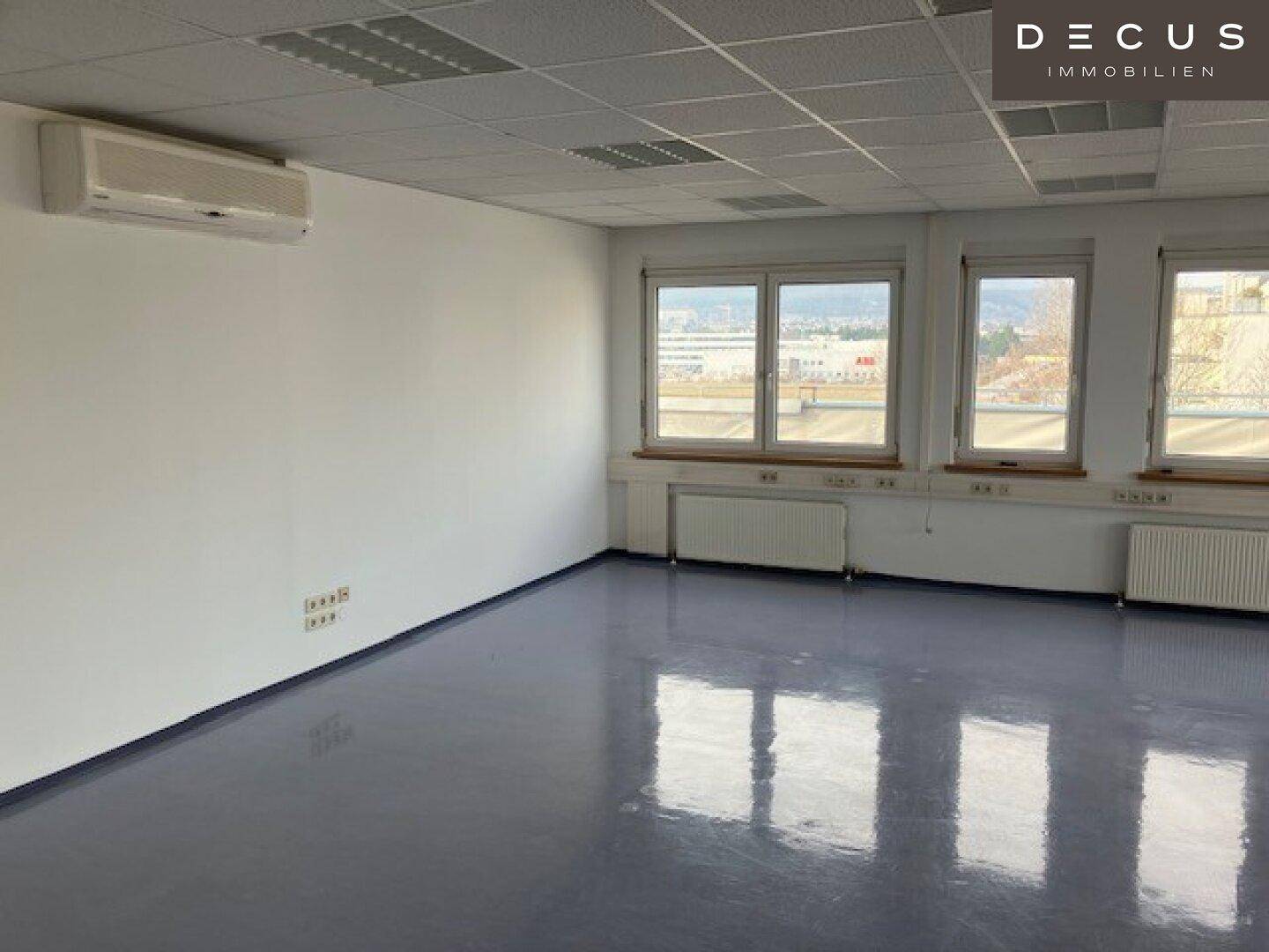 Leeres Büro mit Klimaanlage, hellgrauem Vinylboden und Fenstern mit Blick nach draußen.