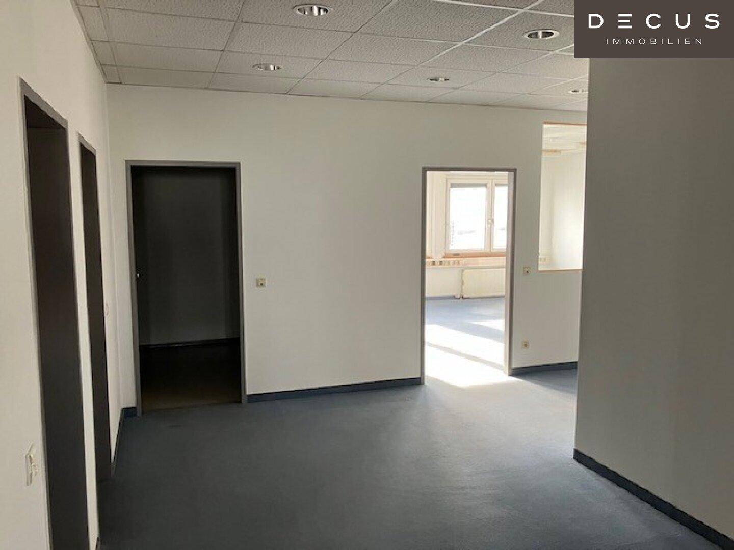 Flur mit mehreren Türen zu Büroräumen und Blick auf ein helles Zimmer mit Fenstern.