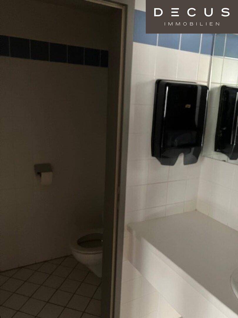 Toilettenbereich mit Fliesenboden und Wandspiegel, Teil eines größeren Waschraums.