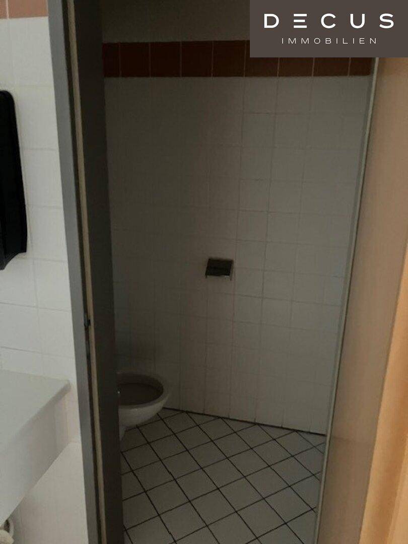 Kleiner Toilettenraum mit weißen Fliesen, braunem Akzentstreifen und einfacher Ausstattung.