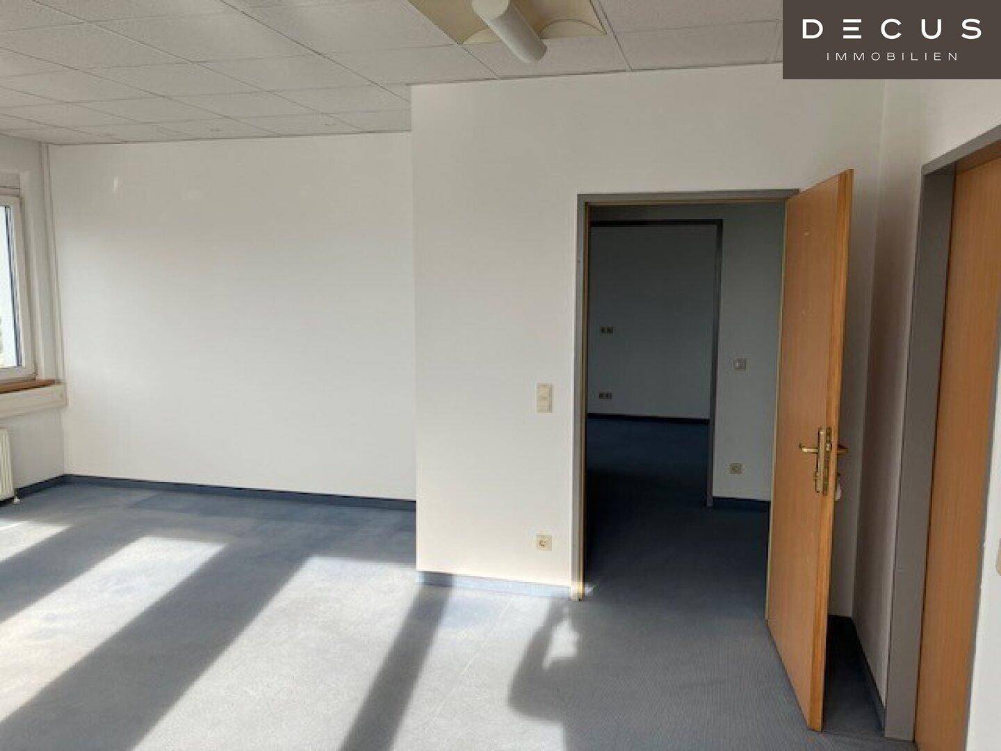 Geräumiger Büroraum mit hellem Boden und Zugang zu einem weiteren Zimmer, gut beleuchtet.