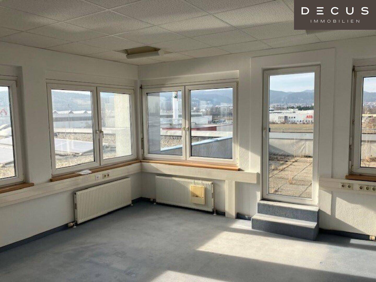 Heller Büroraum mit großen Fenstern und Zugang zu einem Außenbereich, bietet weite Aussicht.