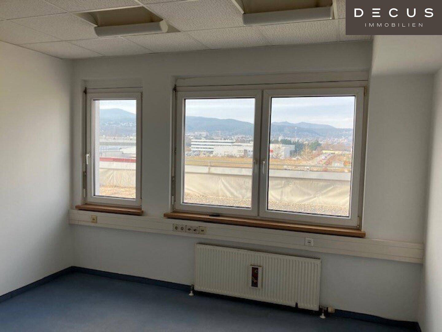 Büroraum mit großen Fenstern und Blick auf die umliegende Landschaft und Gebäude.
