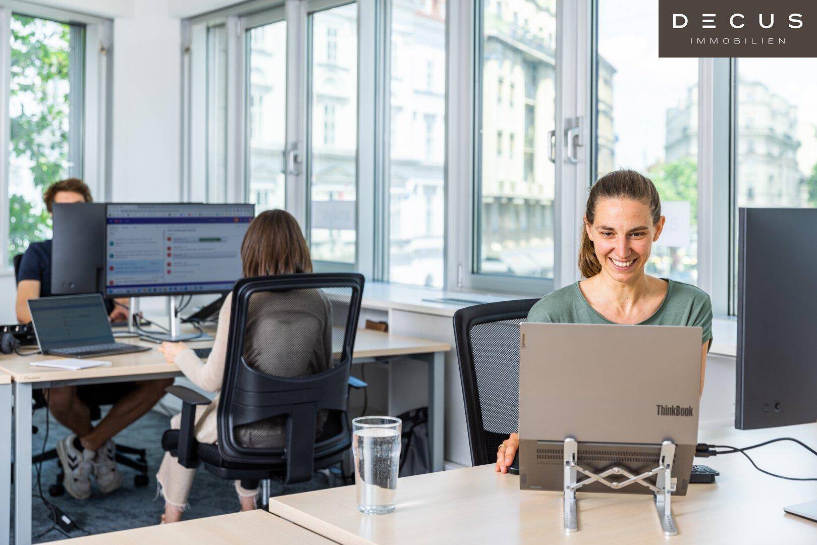 Mitarbeiter arbeiten konzentriert in einem hellen Büro mit großen Fenstern und Blick auf die Stadt.