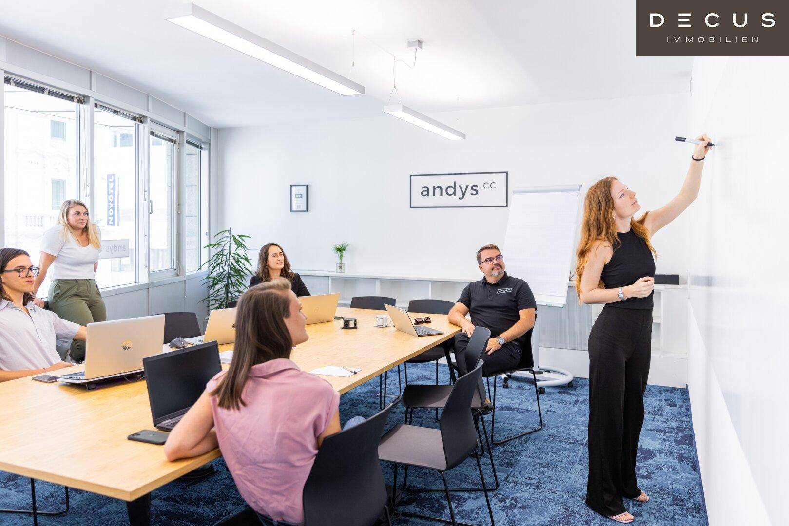 Besprechungsraum mit großem Holztisch, Stühlen, Whiteboard und Mitarbeitern bei einer Präsentation.